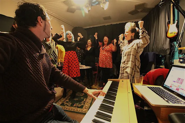 A group of singers with raised fists singing together around a piano at a Discover & Design Your Authentic Voice workshop in Bristol